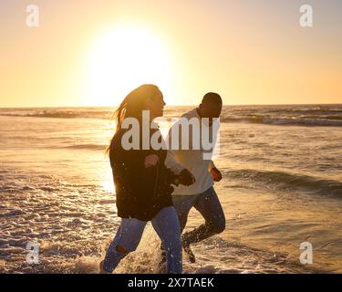 Lässig Gekleidetes, Liebevolles Junges Paar, Das Bei Sonnenaufgang Entlang Der Beach Shoreline Durch Die Wellen Läuft Stockfoto