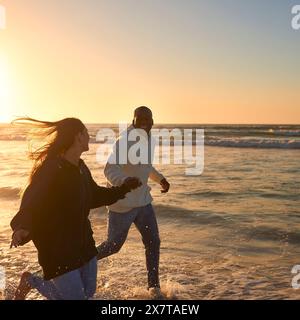 Lässig Gekleidetes, Liebevolles Junges Paar, Das Bei Sonnenaufgang Entlang Der Beach Shoreline Durch Die Wellen Läuft Stockfoto