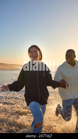 Lässig Gekleidetes, Liebevolles Junges Paar, Das Bei Sonnenaufgang Entlang Der Beach Shoreline Durch Die Wellen Läuft Stockfoto