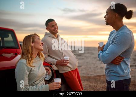 Gruppe Von Freunden, Die Sich Im Auto Mit Heißem Getränk Am Strand Unterhalten Und Gemeinsam Den Sonnenaufgang Beobachten Stockfoto