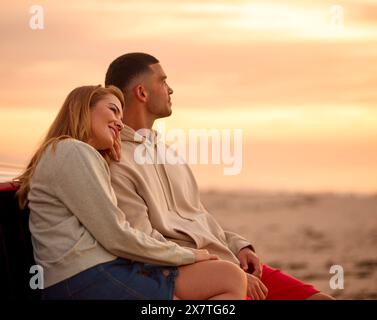Liebevolles Junges Paar, Das Auf Dem Fender Of Car Am Strand Sitzt Und Gemeinsam Den Sonnenaufgang Beobachtet Stockfoto
