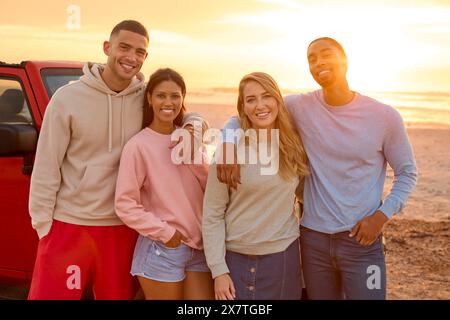 Porträt Eines Paares Mit Freunden, Die Am Strand Mit Dem Auto Stehen Und Gemeinsam Den Sonnenaufgang Beobachten Stockfoto