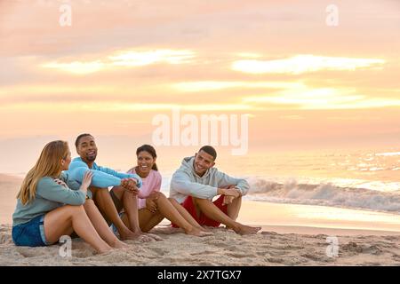 Junges Paar Mit Freunden In Lässiger Kleidung Im Urlaub Sitzen Am Strand Und Beobachten Gemeinsam Den Sonnenaufgang Stockfoto