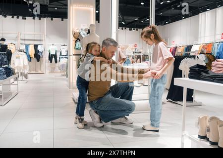 Ein liebevoller Vater hilft seinen Töchtern beim Einkaufen von Kleidung in einem hell erleuchteten Laden Stockfoto