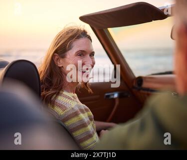 Pensioniertes Seniorenpaar Im Urlaub, Das Im Klassischen Sportwagen Am Strand Sitzt Und Den Sonnenaufgang Beobachtet Stockfoto