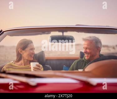 Seniorenpaar Im Ruhestand In Einem Klassischen Sportwagen Mit Heißem Getränk Am Strand, Der Sonnenaufgang Beobachtet Stockfoto