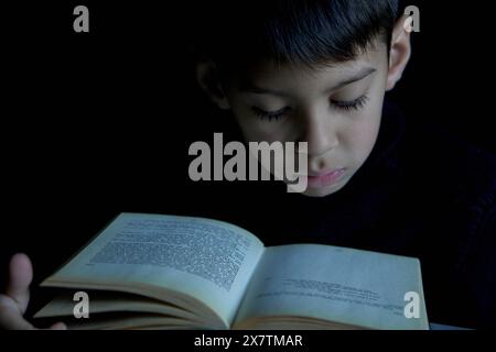 Nahporträt eines intelligenten Latino-Jungen-Gesichts, das in tiefer Konzentration im Dunkeln ein Buch liest. Das Konzept des begabten Kindes Stockfoto