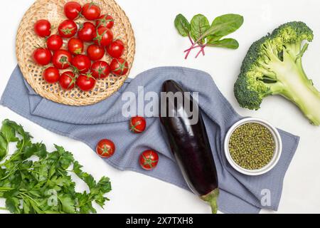 Kirschtomaten in Korbplatte, Auberginen und Mungbohnen in Schüssel auf grauer Serviette. Brokkoli, Mangold-Blätter und Petersilienzweige auf dem Tisch. Weißer Hintergrund. Stockfoto