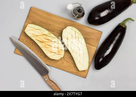 Auberginen-Hälften auf Schneidebrett. Ganze Auberginen, Serviette und Soße auf dem Tisch. Grauer Hintergrund. Flache Lagen. Stockfoto