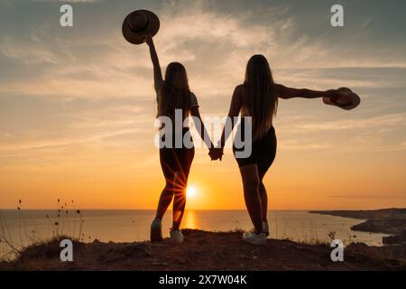 Zwei Frauen stehen auf einem Hügel, halten Hände und tragen Hüte. Die Sonne untergeht im Hintergrund und schafft eine warme und friedliche Atmosphäre. Stockfoto