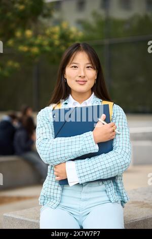 Ein chinesischer Student an der Campus-Universität trägt eine blaue Jacke und Jeans und hält ein blaues Buch Stockfoto