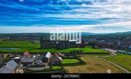 Blick aus der Vogelperspektive auf eine historische Abteiruine auf einem Hügel mit Blick auf eine kleine Stadt mit grünen Feldern und Häusern. Der Himmel ist teilweise bewölkt mit einer Mischung aus Stockfoto