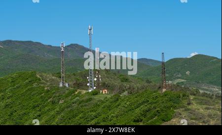 Antennen für Mobilfunknetze befinden sich auf einem grünen Hügel mit Bergen im Hintergrund unter einem klaren blauen Himmel. In der Natur installierte Zellentürme. Stockfoto