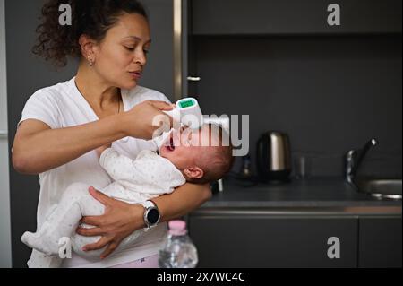 Mutter misst die Körpertemperatur ihres Babys mit einem kontaktlosen elektronischen Infrarot-Thermometer. Kinderbetreuung. Integritätsprüfung. Babyhaube. Hook Stockfoto