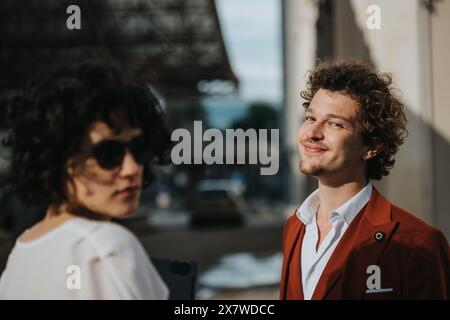 Stilvoller junger Mann, der grinst, während eine Frau mit lockigem Haar und Sonnenbrille draußen wegschaut Stockfoto