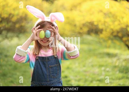 Osterfeier. Ein kleines Mädchen in Hasenohren, das die Augen mit bemalten Eiern im Freien bedeckt, Platz für Text Stockfoto