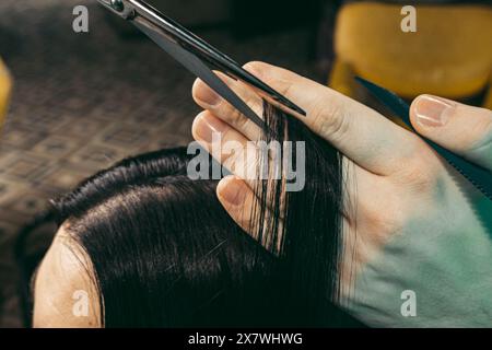 Der Friseur macht eine Frisur für Frauen. Mann Friseur mit einem langen Bart in einer Weste, macht gekonnt ein Friseurstyling mit einem Kamm und einer Haarschneideschere zu einer Frau mit langen schwarzen Haaren in einem Friseurgeschäft. Konzept der Fähigkeiten Kundenansatz.Nahaufnahme Schuss Stockfoto