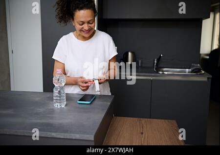 Lächelnde hübsche Frau in weißem T-Shirt mit moderner, intelligenter Armbanduhr mit schwarzem Digitalbildschirm, die im minimalistischen Stil an der Küchenzeile steht Stockfoto