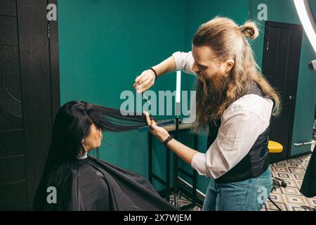 Der Friseur macht eine Frisur für Frauen. Mann Friseur mit langem Bart in einer Weste, macht gekonnt ein Friseurstyling mit einem Kamm und einer Haarschneideschere zu einer Frau mit langen schwarzen Haaren in einem Friseurgeschäft. Konzept der Fähigkeiten Kundenansatz. Mittlere Aufnahme Stockfoto