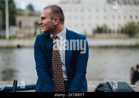 Selbstbewusster Geschäftsmann in blauem Anzug, der draußen am Wasser sitzt Stockfoto