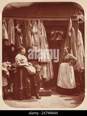 Ein Alter Kleiderladen, Sieben Zifferblätter. Vintage London Street Photography, Ende des 19. Jahrhunderts, von John Thomson, 1877. Das Bild zeigt ein Bekleidungsgeschäft mit zwei Frauen und einem Kind. Stockfoto