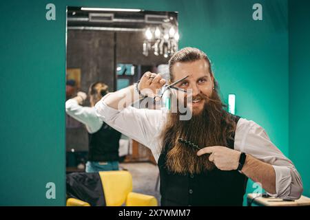 Mann Barbier Porträt. Männlicher europäischer junger Friseur fröhlich bärtig langhaarig, stehend mit Friseurschere in Keksen und lächelnd im Schönheitssalon Friseur, Porträt. Concept Professional Friseur Stockfoto