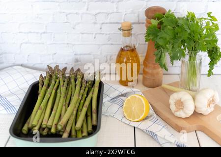 Knuspriger grüner Spargel in Backblech gelegt, zum Kochen vorbereitet. Abgebildet an einer Ziegelwand, einer Olivenflasche und anderen Zutaten Stockfoto