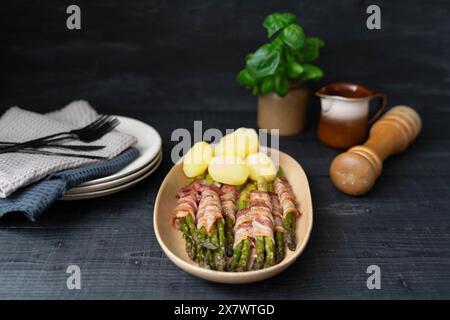 Gekochter grüner Spargel in Speck gewickelt und mit gekochten Kartoffeln auf schönen beigen Tellern auf dunklem Hintergrund serviert Stockfoto