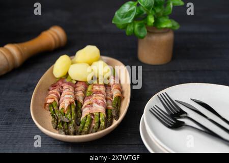 Gekochter grüner Spargel, gebacken in Speck und serviert mit gekochten Kartoffeln auf schönen beigen Tellern auf dunklem Hintergrund Stockfoto