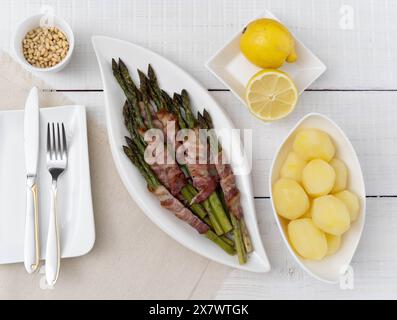 Grüner Spargel, gebacken in Speck und serviert mit gekochten Kartoffeln auf schönen weißen Tellern auf hellem Holz Stockfoto