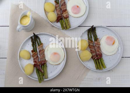 Grüner Spargel, gebacken in Speck und serviert mit gekochten Kartoffeln und Eiern, auf schönen weißen Tellern auf hellem Holz Stockfoto