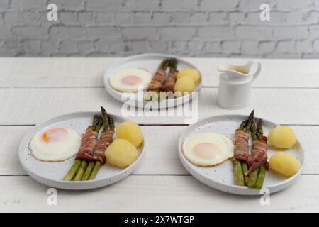 Grüner Spargel, gebacken in Speck und serviert mit gekochten Kartoffeln und Eiern, auf schönen weißen Tellern auf hellem Holz Stockfoto