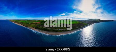 Aus der Vogelperspektive auf einen schmalen Küstenstreifen mit üppigen grünen Feldern auf der einen Seite und dem Meer auf der anderen unter einem klaren blauen Himmel. Stockfoto