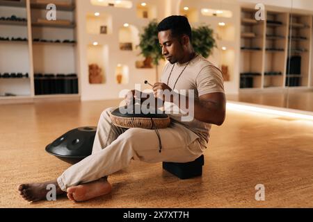 Glücklicher ruhiger schwarzer Schlagzeuger, der in Trance spielt, mit Drumsticks auf Glucophon, der im Kreuzbein auf dem Boden im gemütlichen Meditationsraum sitzt. Stockfoto