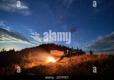 Camping in den Bergen unter bewölktem Nachthimmel. Junger Mann, der sich am Lagerfeuer ausruht, in der Nähe eines Touristenzelts und eines Geländewagens auf einem Hügel. Ein reisender Mann, der nachts die Sterne beobachtet. Stockfoto