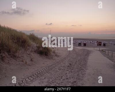 Ein friedlicher Pfad führt durch die Sanddünen zum Strand bei einem orangen Sonnenuntergang, untergehende Sonne an einem Strand mit Liegestühlen und Wolken am Himmel, spi Stockfoto