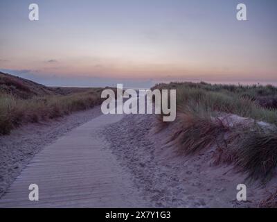 Holzweg durch Sanddünen bei Sonnenuntergang, führt zum Meer unter einem Dämmerungshimmel, Sonnenuntergang an einem Strand mit Liegestühlen und Wolken am Himmel, spi Stockfoto
