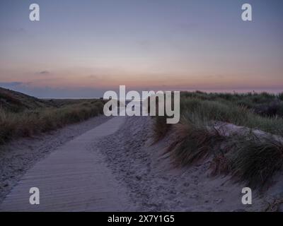 Dämmerungsszene mit Holzweg durch Dünen, der bei Sonnenuntergang zum Meer führt, untergehende Sonne an einem Strand mit Liegestühlen und Wolken am Himmel, Spiekeroo Stockfoto