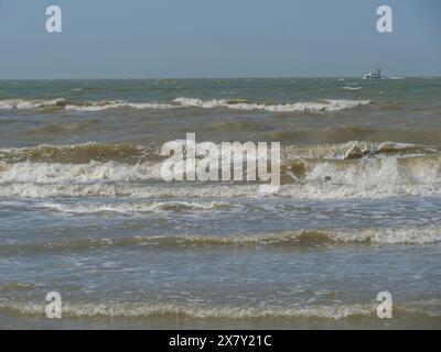 Stürmisches Meer mit starken Wellen und einem Boot am Horizont, breiter Sandstrand am Meer mit Möwen und einem Stadtpanorama auf der Promenade, de haan, Belgi Stockfoto