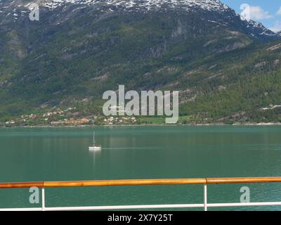 Ein einsames Segelboot gleitet über den ruhigen, grünen Fjord, umgeben von dramatischen Bergen und üppiger Vegetation, und segelt in einem Fjord mit grünem Wasser und sno Stockfoto