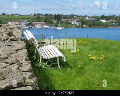 Zwei weiße Bänke an einer Steinmauer, Gras davor und ein See mit Booten und Häusern dahinter, teilweise bewölkter Himmel, alte Steinkirche und viele Grabsteine an Stockfoto