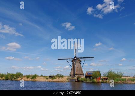 Eine Windmühle neben einem kleinen Haus unter klarem Himmel mit verstreuten Wolken, viele historische Windmühlen an einem Fluss inmitten von Feldern, Kinderdijk, Neth Stockfoto