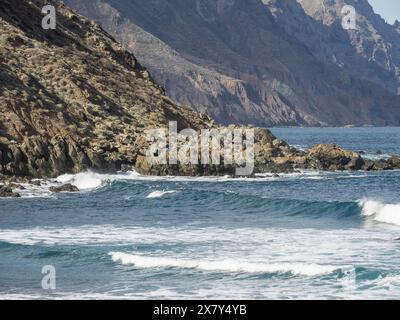 Eine felsige Küstenlandschaft mit Wellen, die gegen die Klippen krachen, Lavastrand mit Bergen und blauem Himmel im Hintergrund, teneriffa, spanien Stockfoto