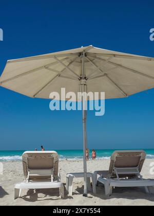 Zwei Liegestühle unter einem großen Sonnenschirm am Strand mit Blick auf das Meer, glitzerndes grünes Meer mit Strand, Sonnenschirmen und Liegen vor einem hellen Blau Stockfoto