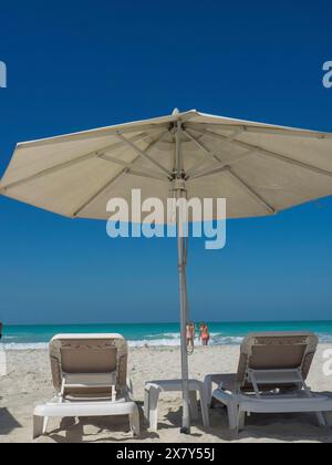 Zwei Liegestühle unter einem großen Sonnenschirm am Strand mit Blick auf das Meer, glitzerndes grünes Meer mit Strand, Sonnenschirmen und Liegen vor einem hellen Blau Stockfoto