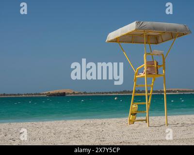 Am Strand steht ein gelber Rettungsschwimmer-Turm mit türkisfarbenem Wasser im Hintergrund und einem klaren blauen Himmel, glitzerndem grünem Meer mit Strand, Sonnenschirmen und Stockfoto