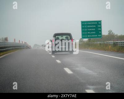 Ein dunkles Auto fährt auf einer regnerischen Autobahn in Italien mit einem Straßenschild, das Wegbeschreibung in die Städte anzeigt, regnerischer Tag auf der Autobahn A1, Piacenza, Italien, Europa Stockfoto