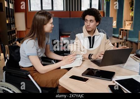 Hochwinkelansicht eines kaukasischen Mädchens mit Behinderung und ihres nahöstlichen Freundes, der in der Schulbibliothek am Tisch sitzt und gemeinsam an der Präsentation arbeitet Stockfoto