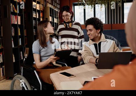 Kaukasisches Mädchen mit Behinderung und ihre asiatischen und nahöstlichen Freunde studieren in der Schulbibliothek mit einem digitalen Tablet und Laptop Stockfoto