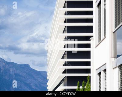 Ein modernes Gebäude mit weißen, horizontalen Platten und geometrischer Fassade, das vor felsigen Bergen unter teilweise bewölktem Himmel liegt. Stockfoto